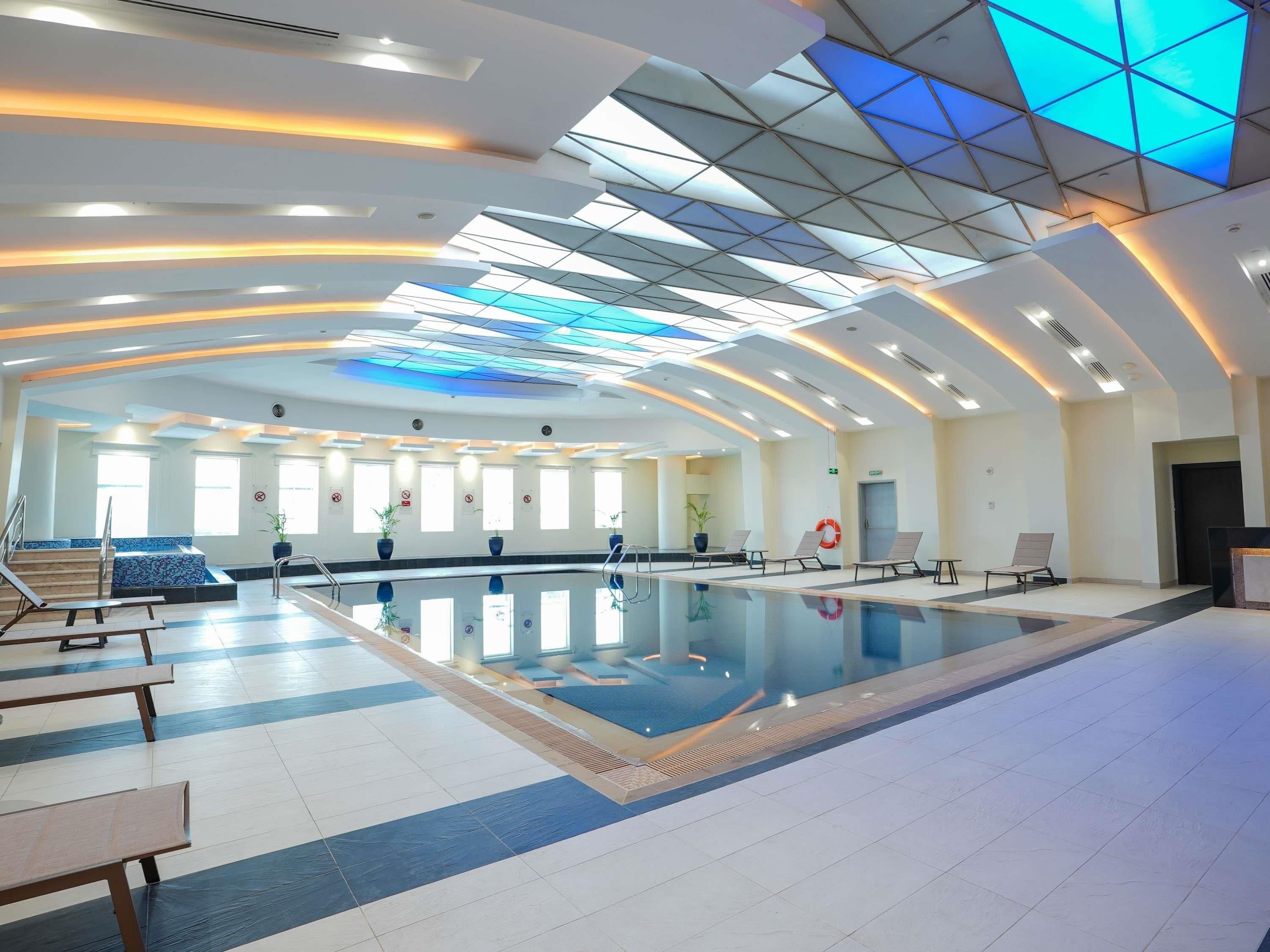 A spacious indoor swimming pool with a modern ceiling design, clear windows, and sunlit areas.