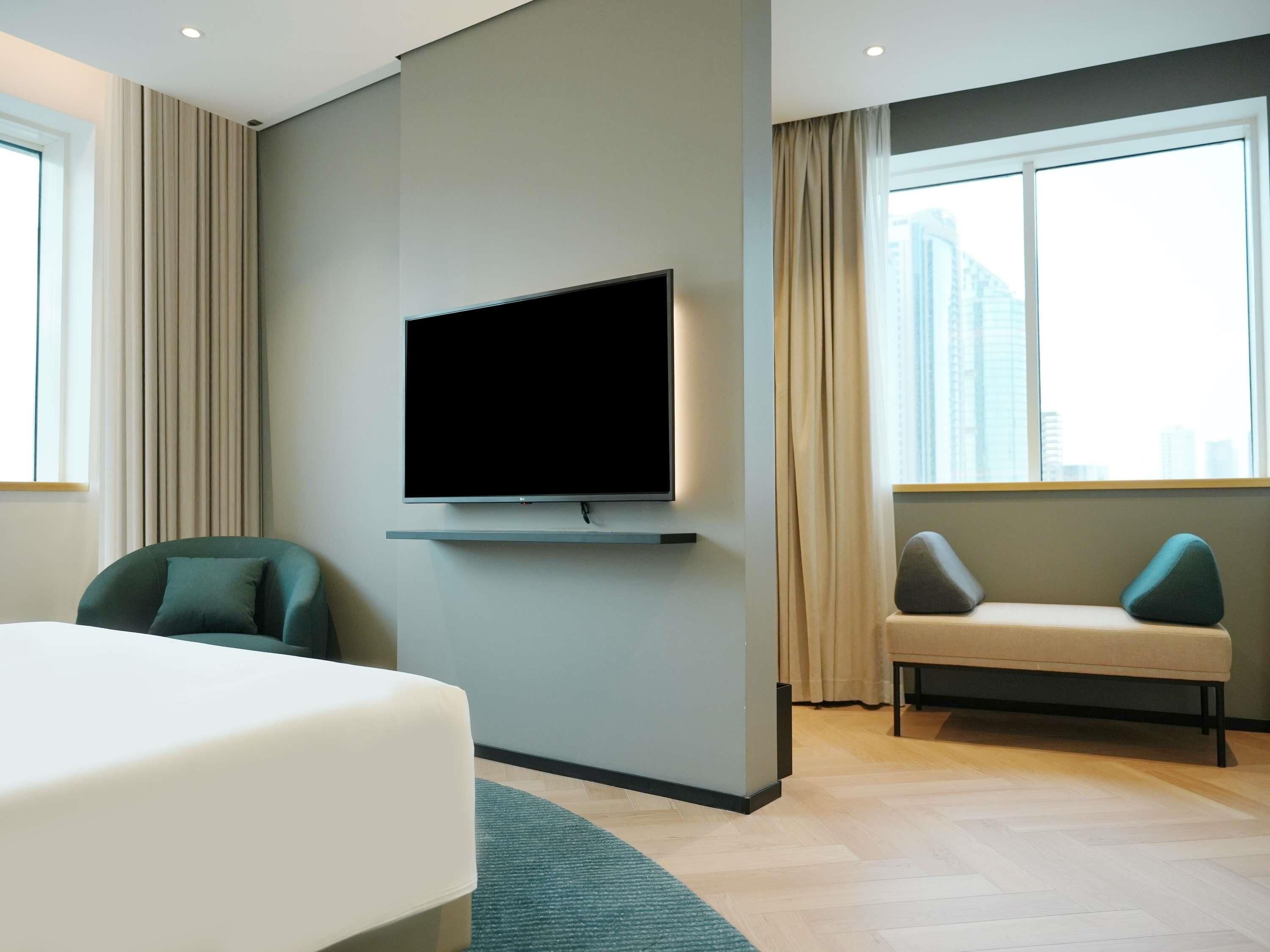 A modern hotel room with a flat-screen TV mounted on the wall, a bed, a seating area with a sofa and armchairs, and large windows offering a city view.