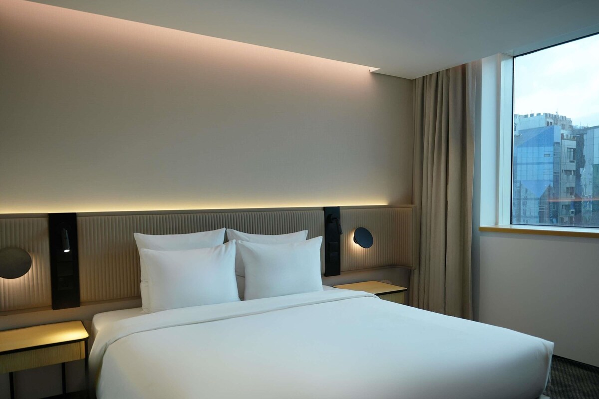 A hotel room with a large bed, two bedside tables, a wall-mounted lamp, and a window with a city view.