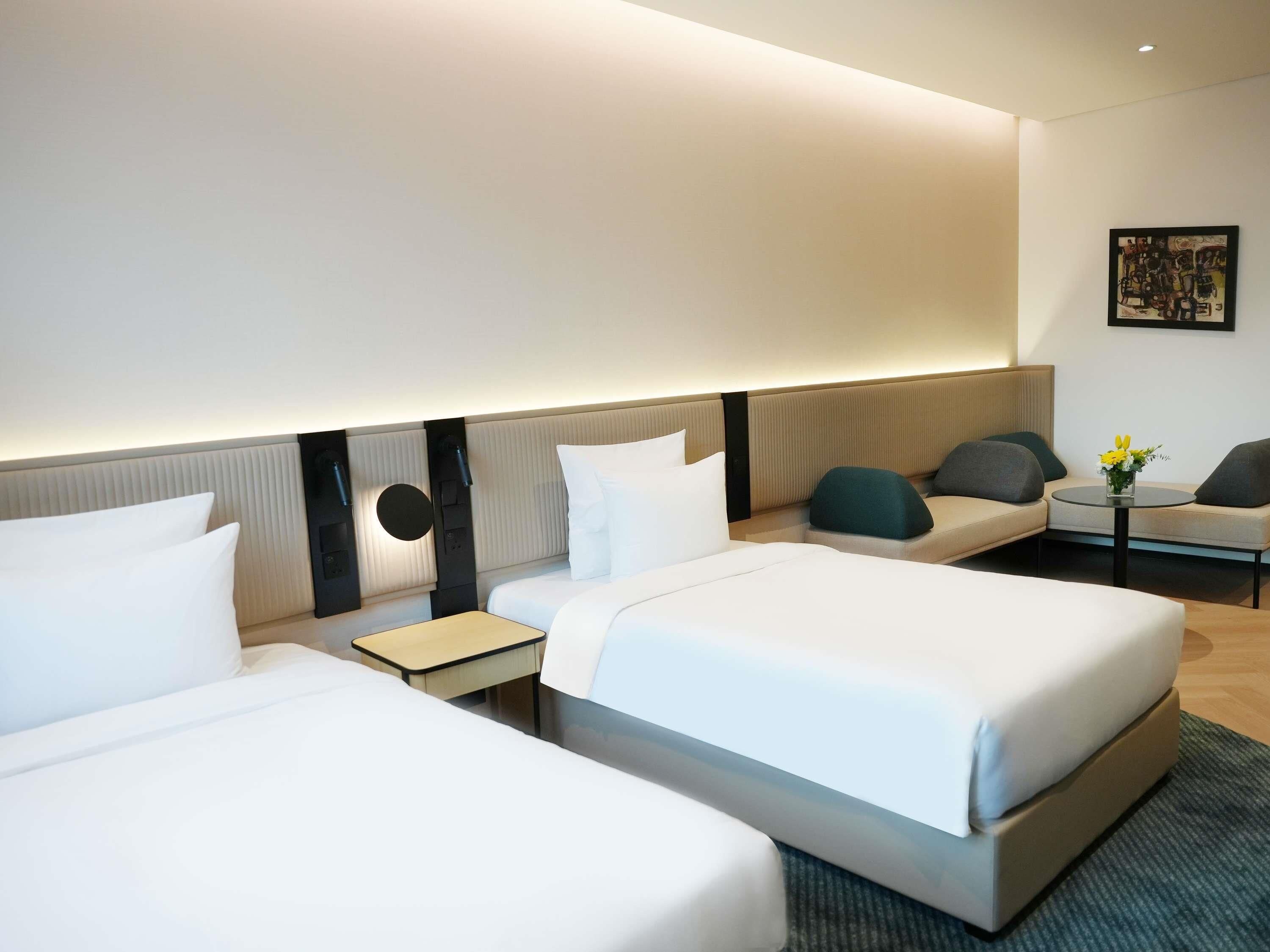 A modern hotel room with two beds, a sofa, a small table with flowers, and a wall-mounted artwork.