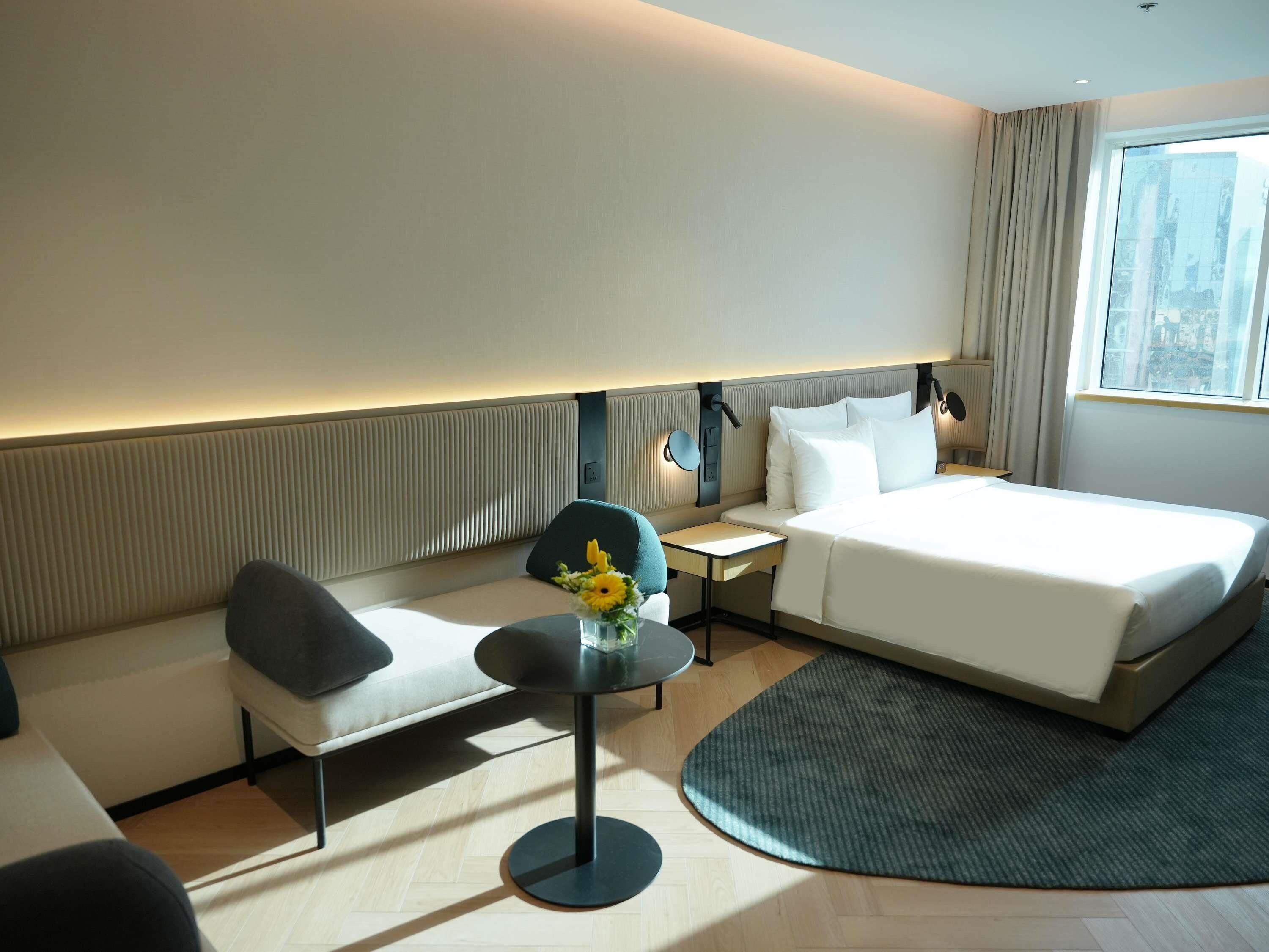 A hotel room with a bed, a seating area with a table, a small round table with a vase, and a view of the city through a large window.