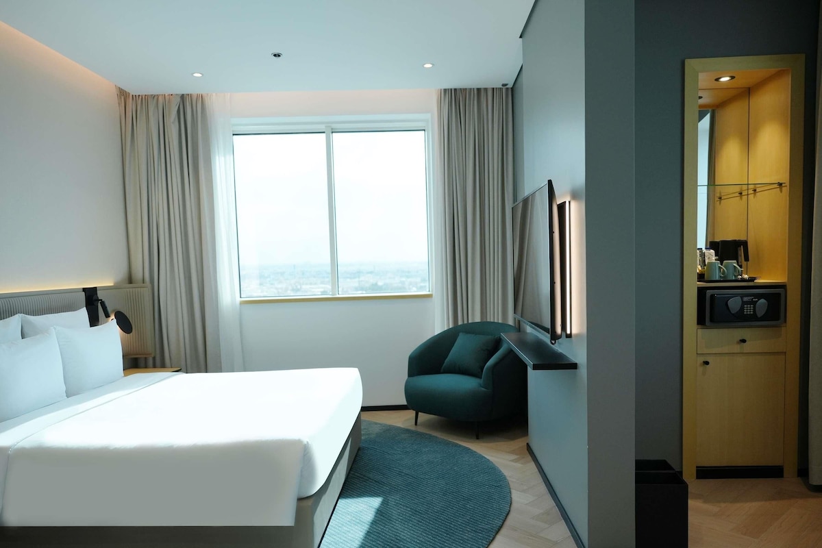 A modern hotel room with a large bed, a desk, a chair, a view of the city, and a minibar.