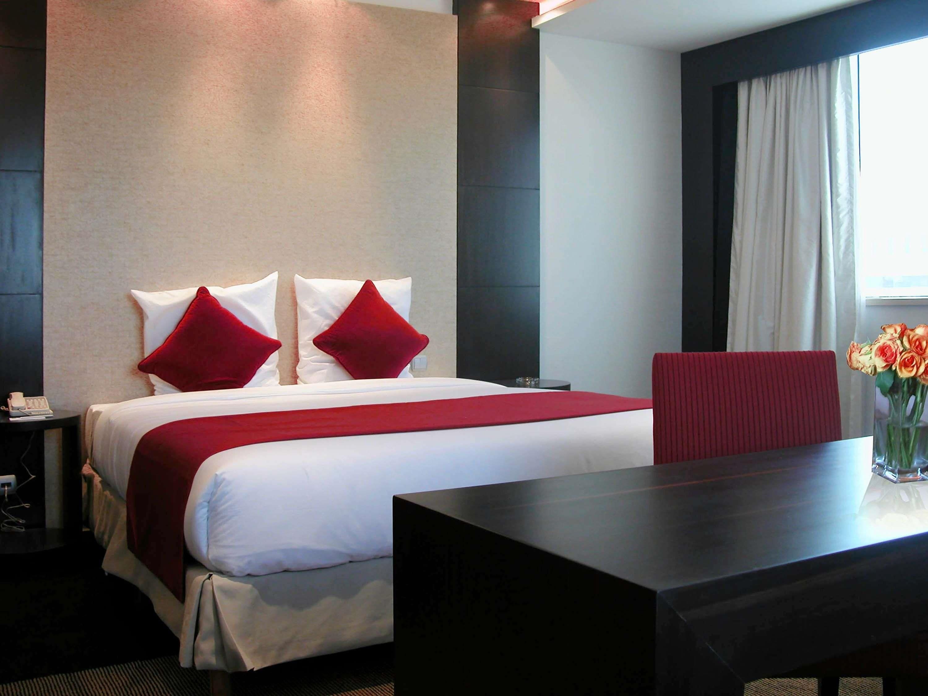 Une chambre d’hôtel avec un lit, une table de chevet, une table ornée d’un vase de fleurs et une fenêtre avec des rideaux.