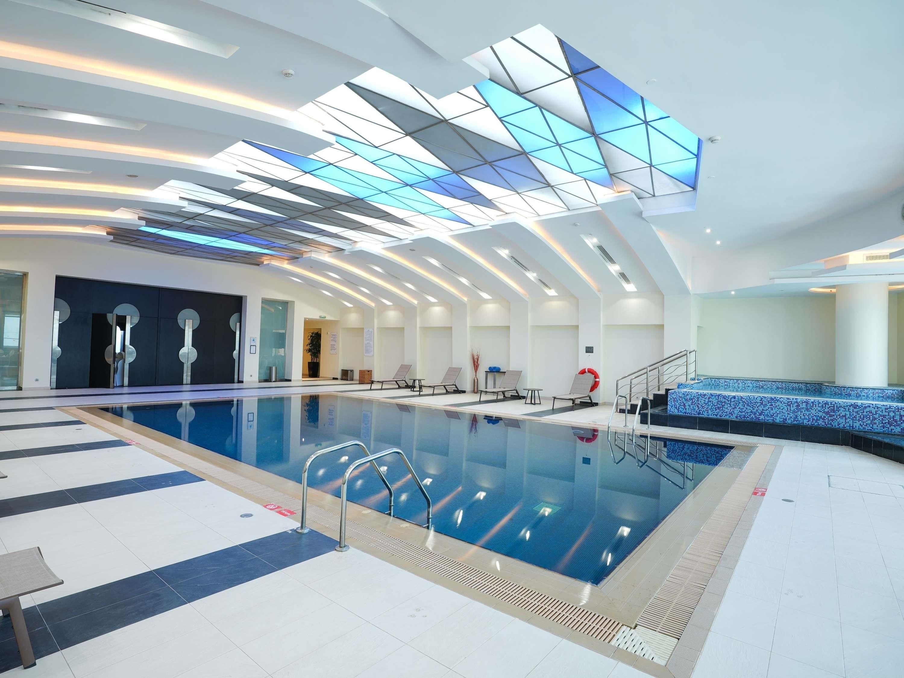 Una piscina interior moderna con techo de vidrio, sillas de descanso y suelo de baldosas.