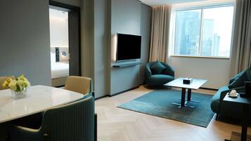 Una habitación de hotel moderna con un televisor de pantalla plana en la pared, un sofá, una mesa de comedor con sillas y vistas a la ciudad a través de grandes ventanales.