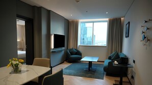 A modern living room with a flat-screen TV, a sofa, a dining table with chairs, and a large window with curtains. - Novotel Riyadh Al Anoud (Riyadh)