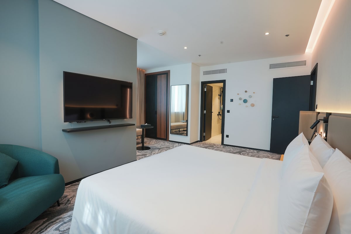 A modern hotel room with a large bed, a flat-screen TV, a seating area, and a bathroom visible through an open door.