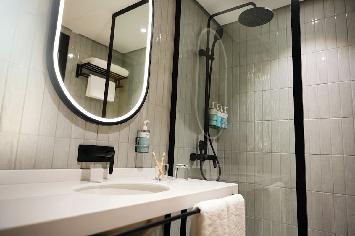 A modern bathroom with a white sink, a large mirror, a handheld showerhead, and a glass of water.