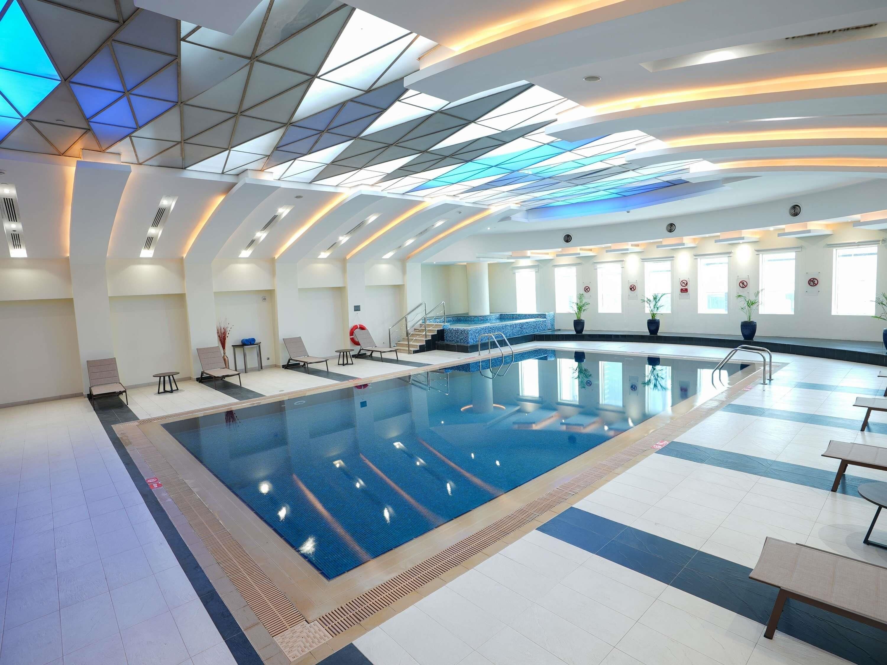 Una piscina interior moderna con un diseño geométrico en el techo, zona de piscina transparente y ventanas iluminadas por el sol.
