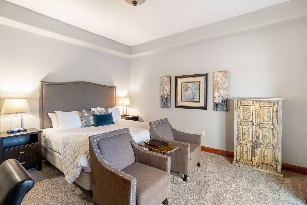 A hotel room with a bed, two armchairs, a small table, a nightstand, and a cabinet.