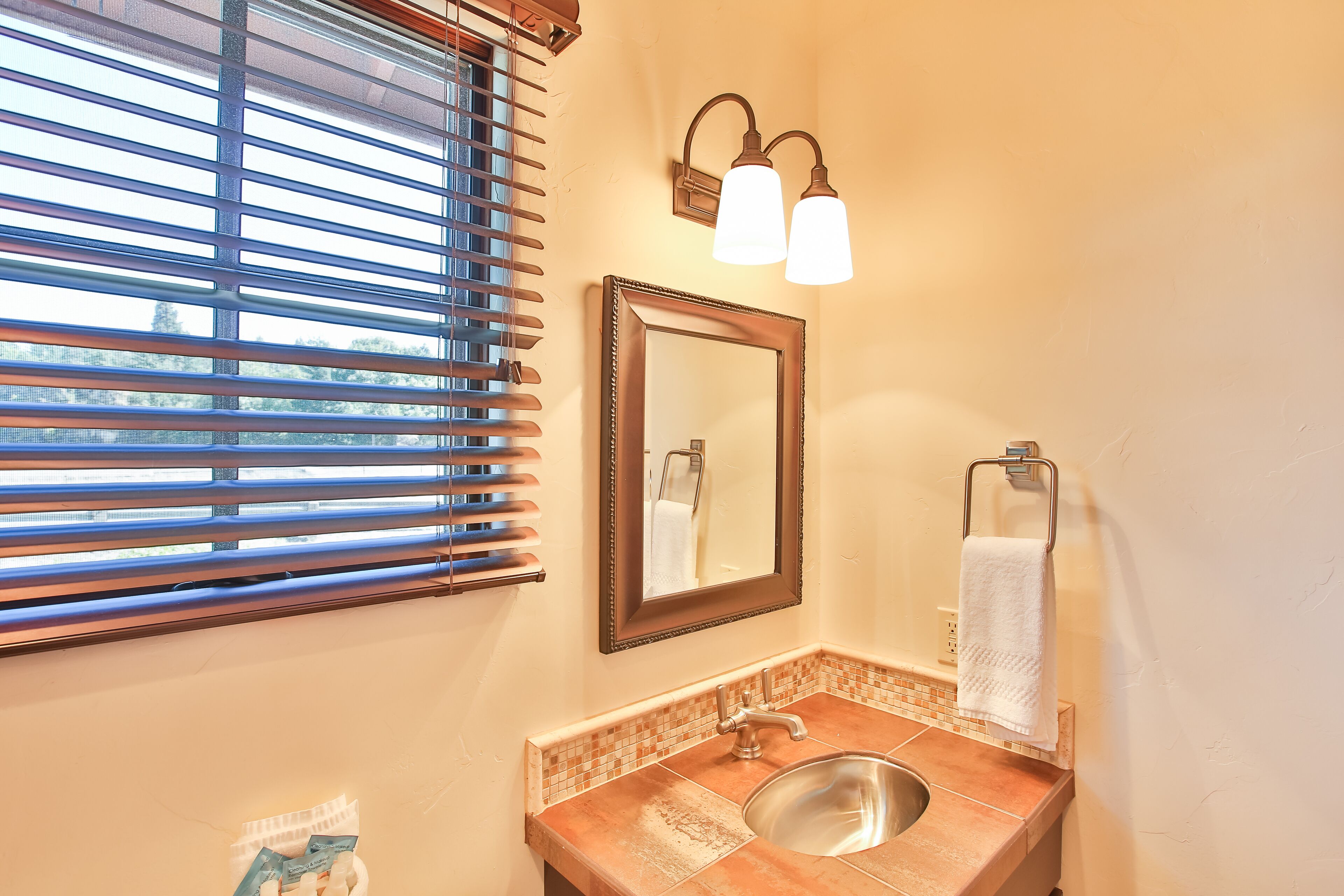 Un baño con lavamanos, espejo, toallero y ventana con persianas.