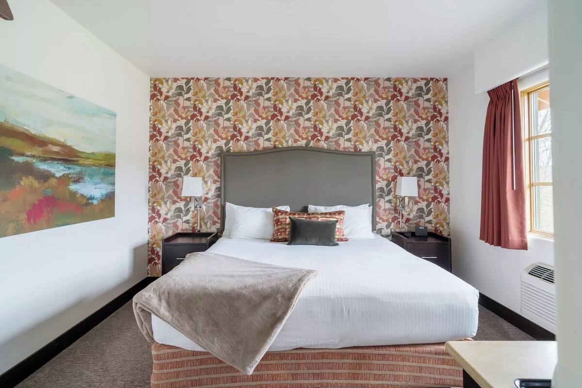 A hotel room with a large bed, floral wallpaper, a painting on the wall, and a window with curtains.
