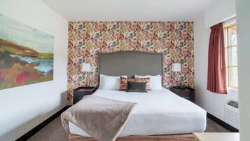 Une chambre d’hôtel avec un grand lit, un papier peint à motifs floraux, un tableau au mur et une fenêtre avec des rideaux.