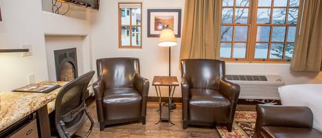 A hotel room with a desk, two leather chairs, a sofa, a TV, a lamp, a window with curtains, and a radiator.