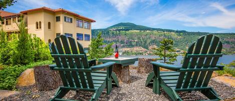 A view of a lakeside property with two Adirondack chairs, a table with a bottle, and a building with a red roof.