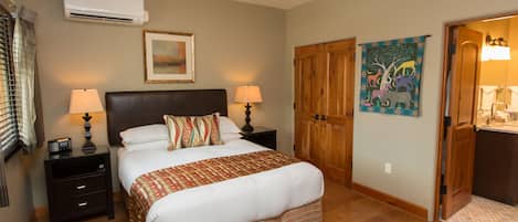 Hotelkamer met een bed, nachtlampen, een nachtkastje, een airconditioner, een houten garderobe, een schilderij en een zichtbare badkamer door een geopende deur.