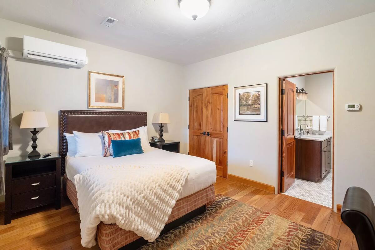A bedroom with a large bed, two bedside tables with lamps, a wooden wardrobe, and a small kitchenette.