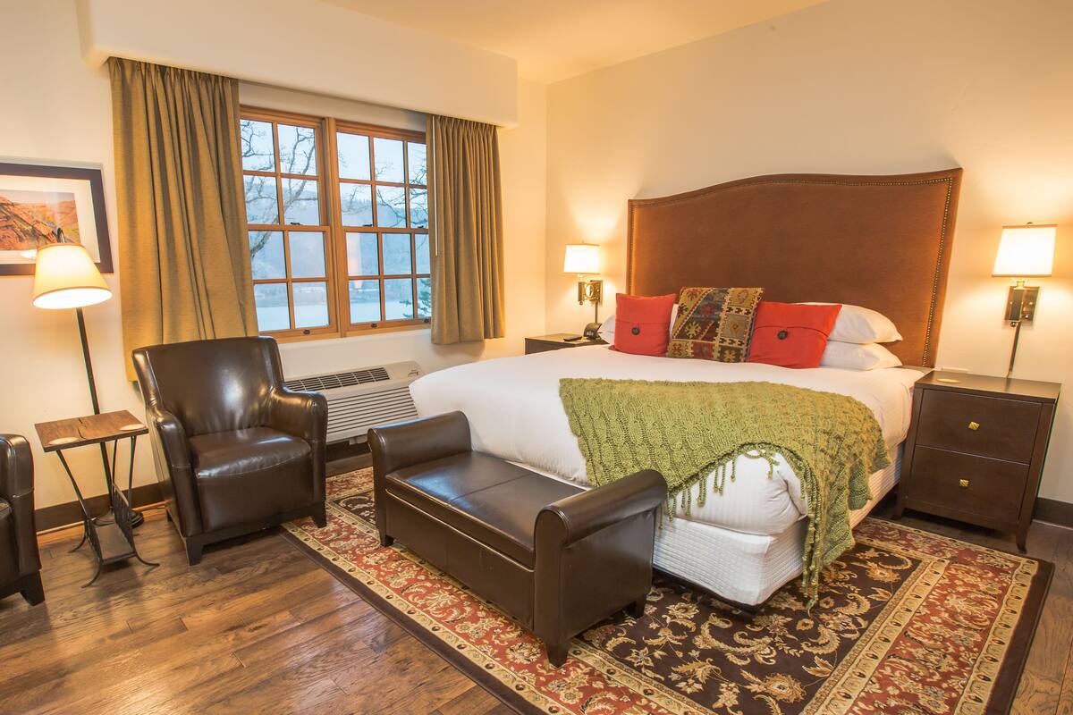 A hotel room with a large bed, a leather armchair, a small sofa, a nightstand with a lamp, and a window with curtains.