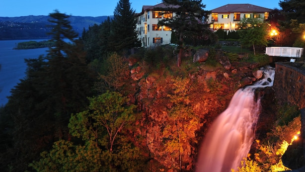 Een meerlaags huis met een waterval ervoor, omringd door bomen en een meer.