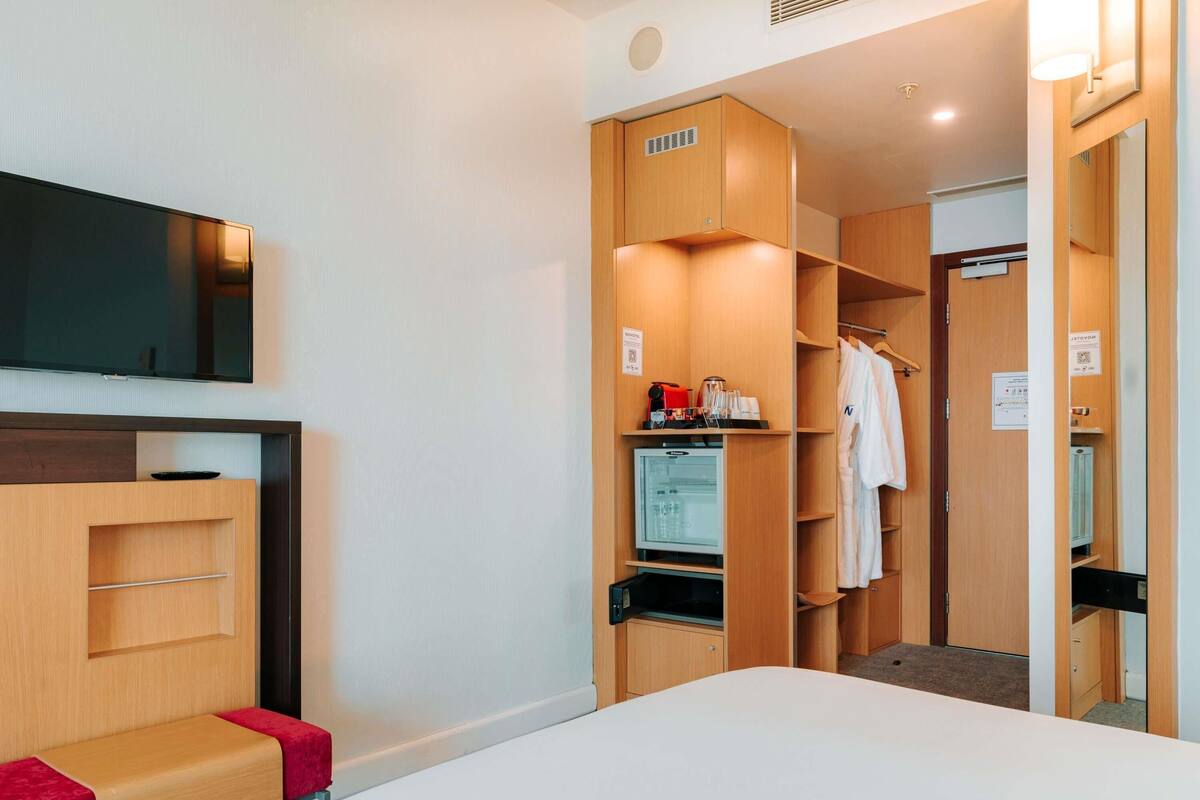 A hotel room with a bed, a TV mounted on the wall, a wardrobe, and a small seating area.