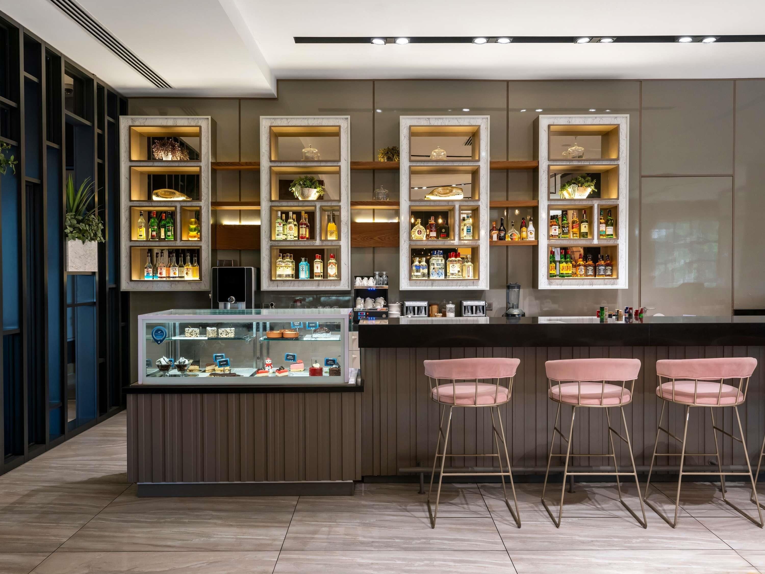 A bar area with shelves of bottles, a display case with desserts, and three pink bar stools.