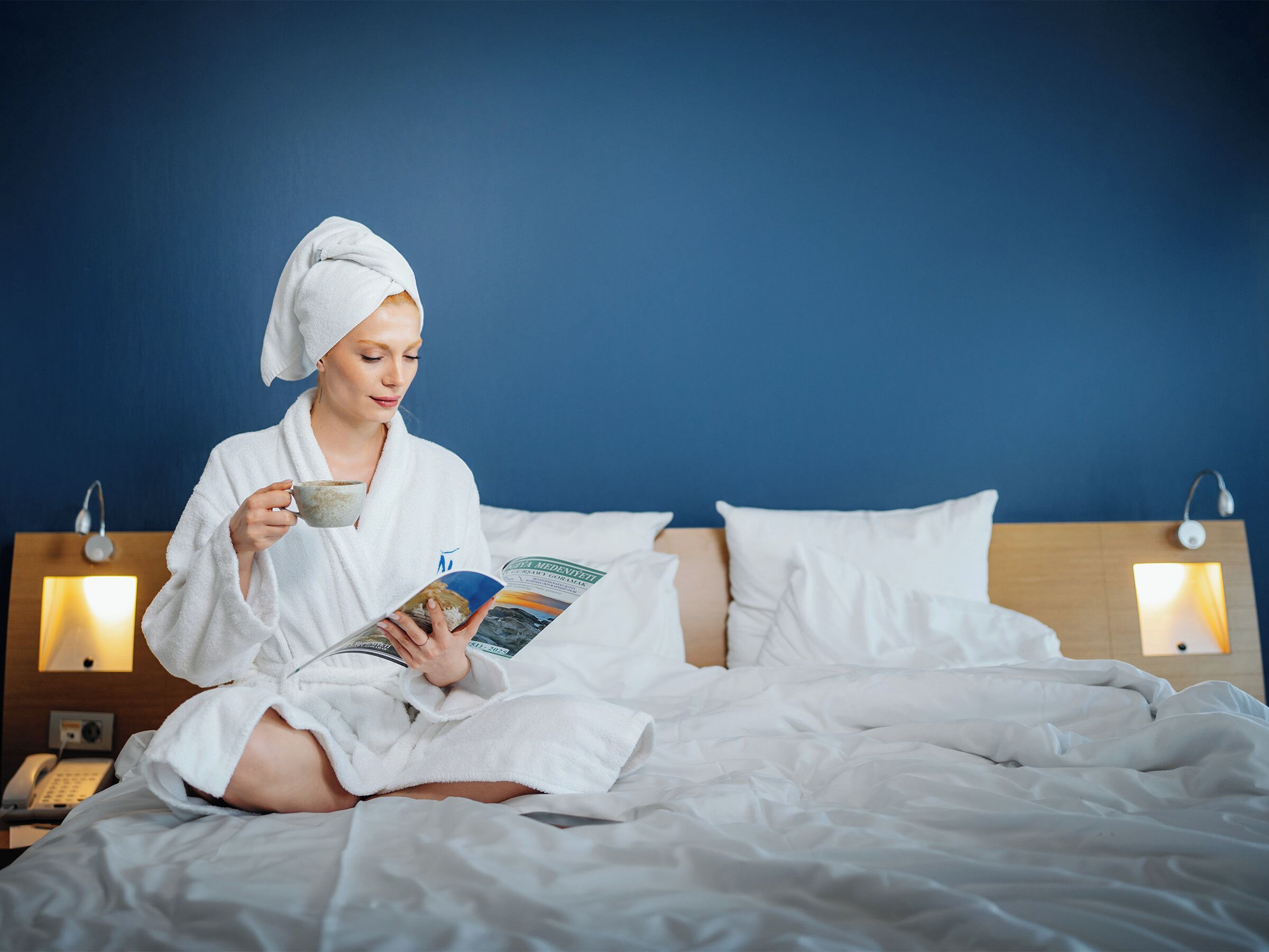 A person in a white bathrobe sitting on a bed with a magazine, a bedside lamp, and a phone on a wooden nightstand.
