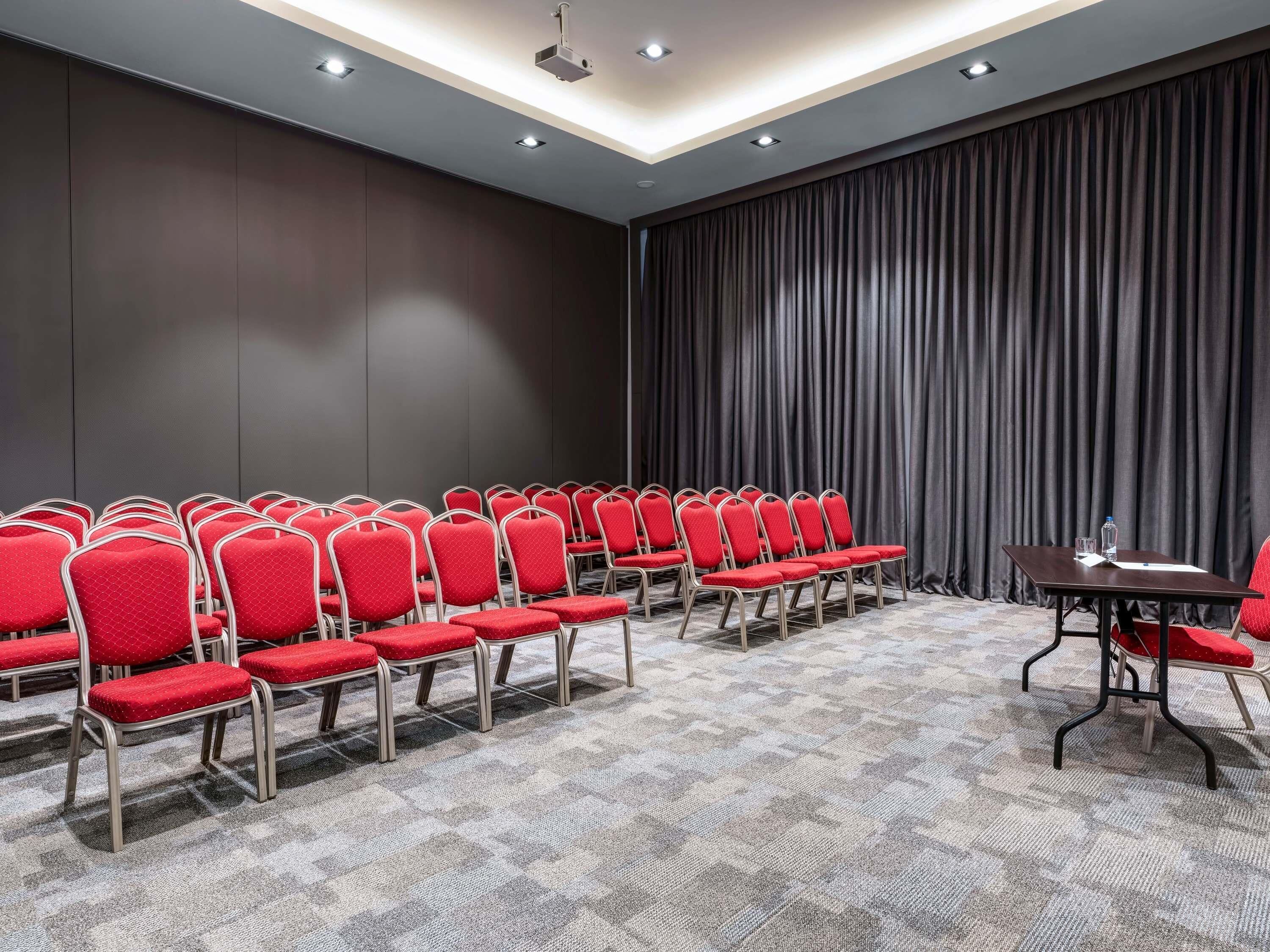 Une salle de conférence avec des rangées de chaises rouges, un fond constitué d’un rideau sombre et un sol recouvert de moquette.