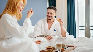 Een koppel in een hotelkamer, beiden in witte badjassen, met een ontbijttafel op het bed.
