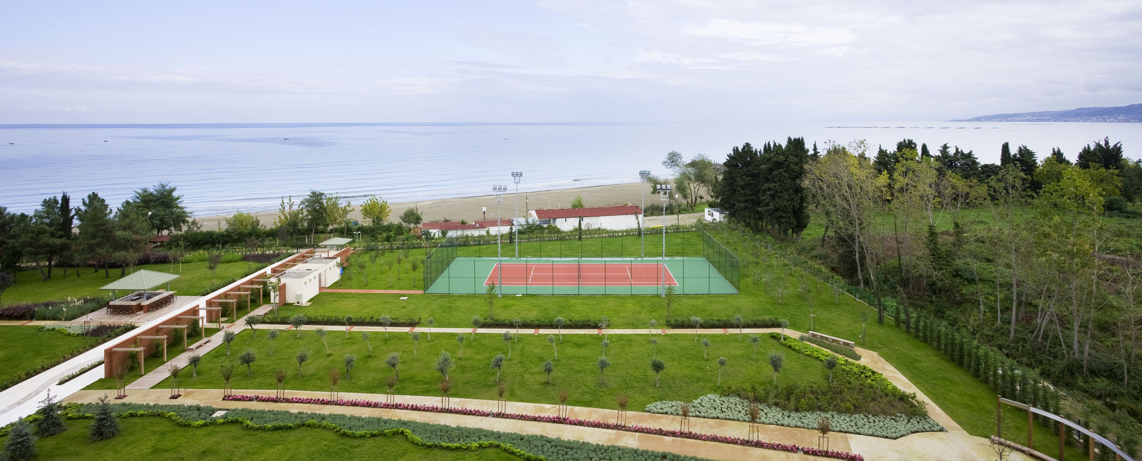 Uma quadra de tênis cercada por gramados verdes e árvores, com vista para o mar ao fundo.