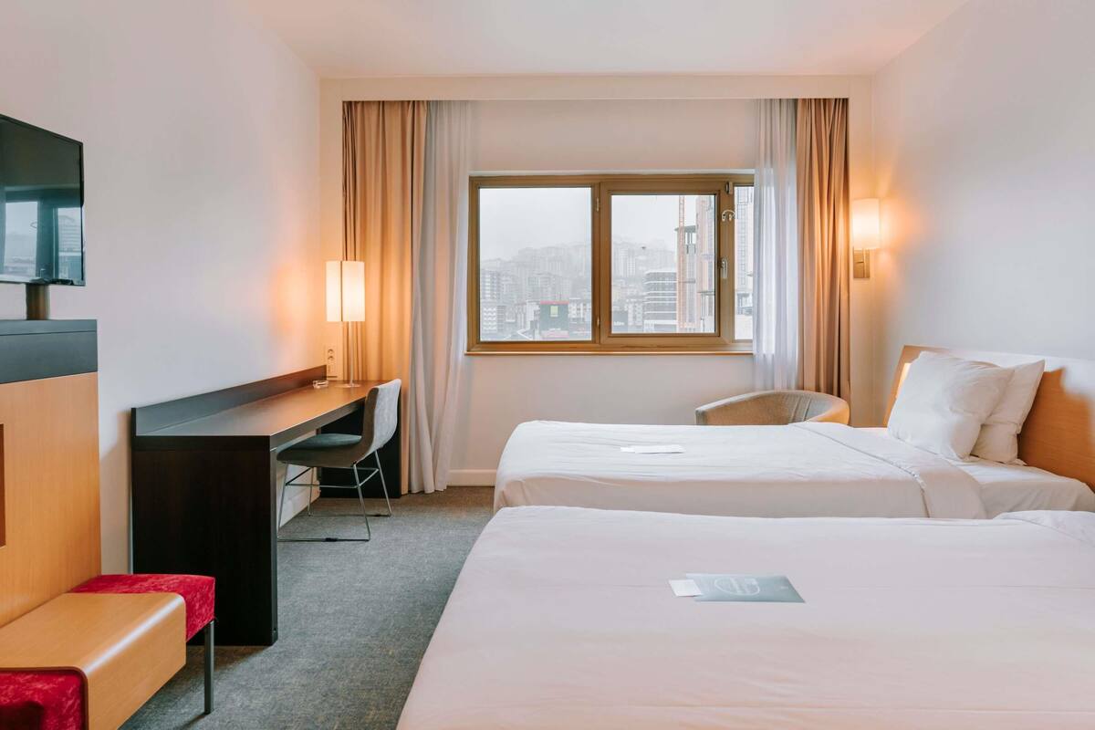 A hotel room with two beds, a desk, a chair, a TV, and a window with a city view.
