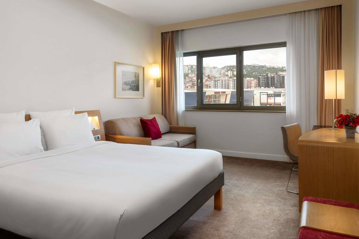 A hotel room with a large bed, a sofa, a desk with a lamp, and a view of the city through the window.
