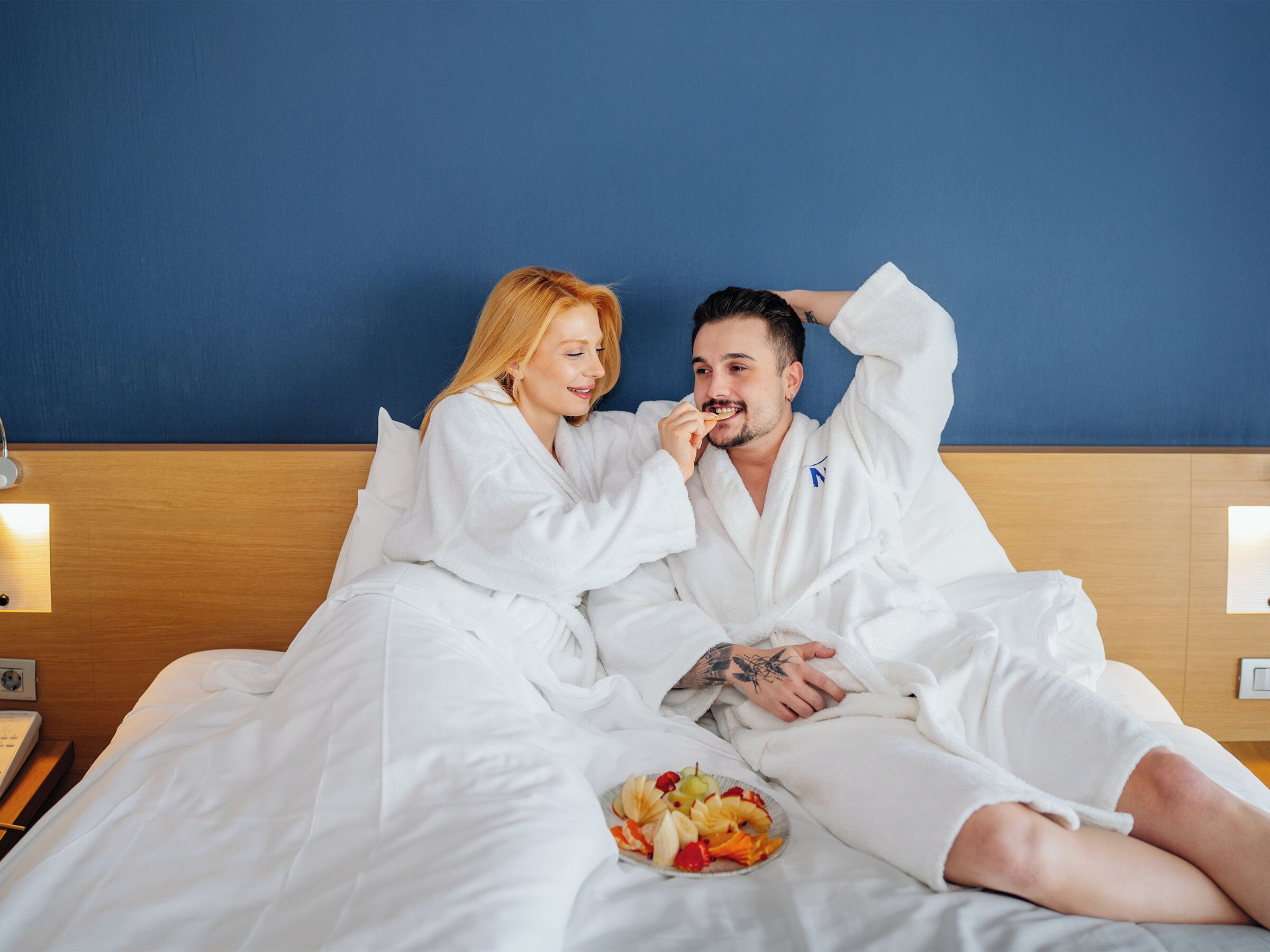 A bed with white bedding, a plate of fruit, and a blue wall in the background.