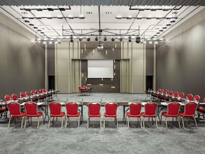 Un salón de conferencias con filas de sillas rojas, una larga mesa, una pantalla de proyector y un diseño moderno en el techo.