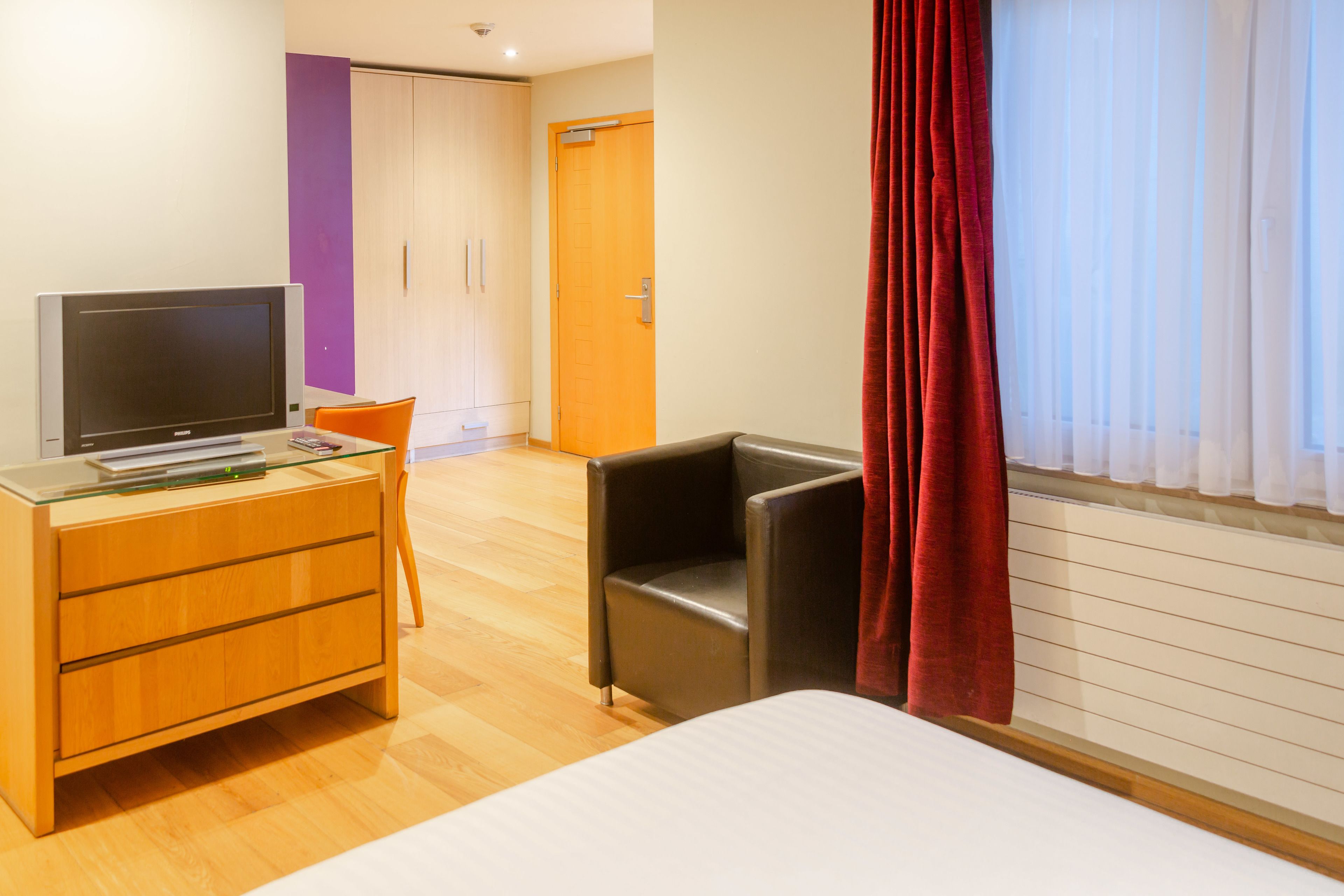 Habitación de hotel con cama, televisor en una cómoda de madera, una silla roja y un armario.