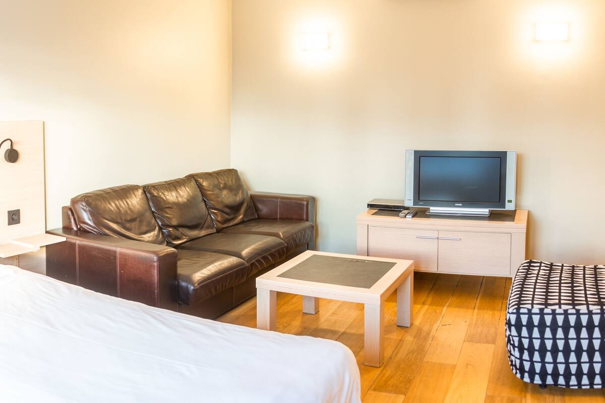 A hotel room with a bed, a leather sofa, a small table, a TV, and a patterned ottoman.