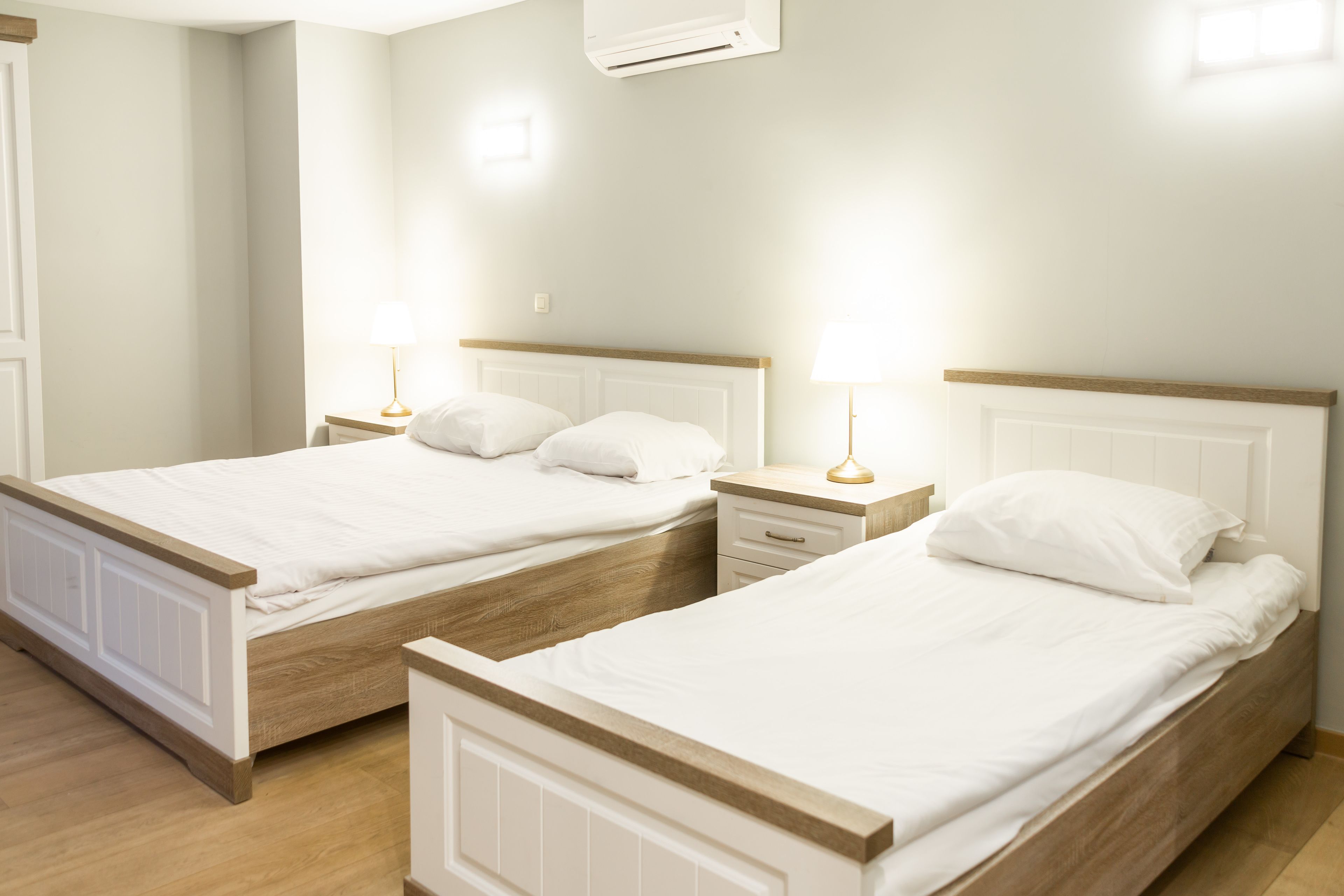 Twee enkele bedden met houten hoofdborden en wit beddengoed, een nachtkastje met een lamp, en een wandairconditioner.