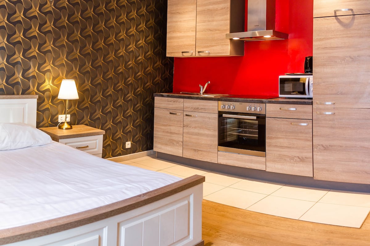 A bedroom with a bed, a nightstand with a lamp, a red wall, and a kitchen area with cabinets and appliances.