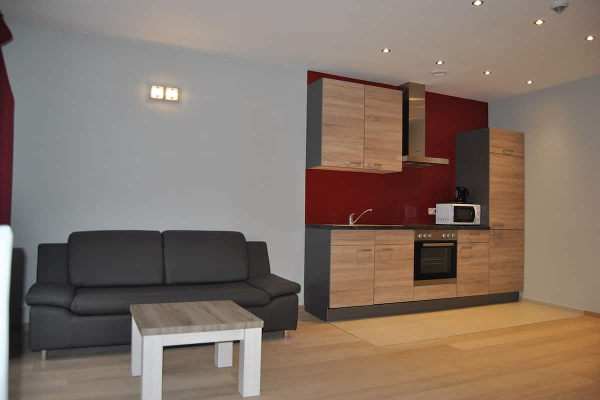 A modern living room with a grey sofa, a small white coffee table, and a kitchen area with wooden cabinets and a microwave.