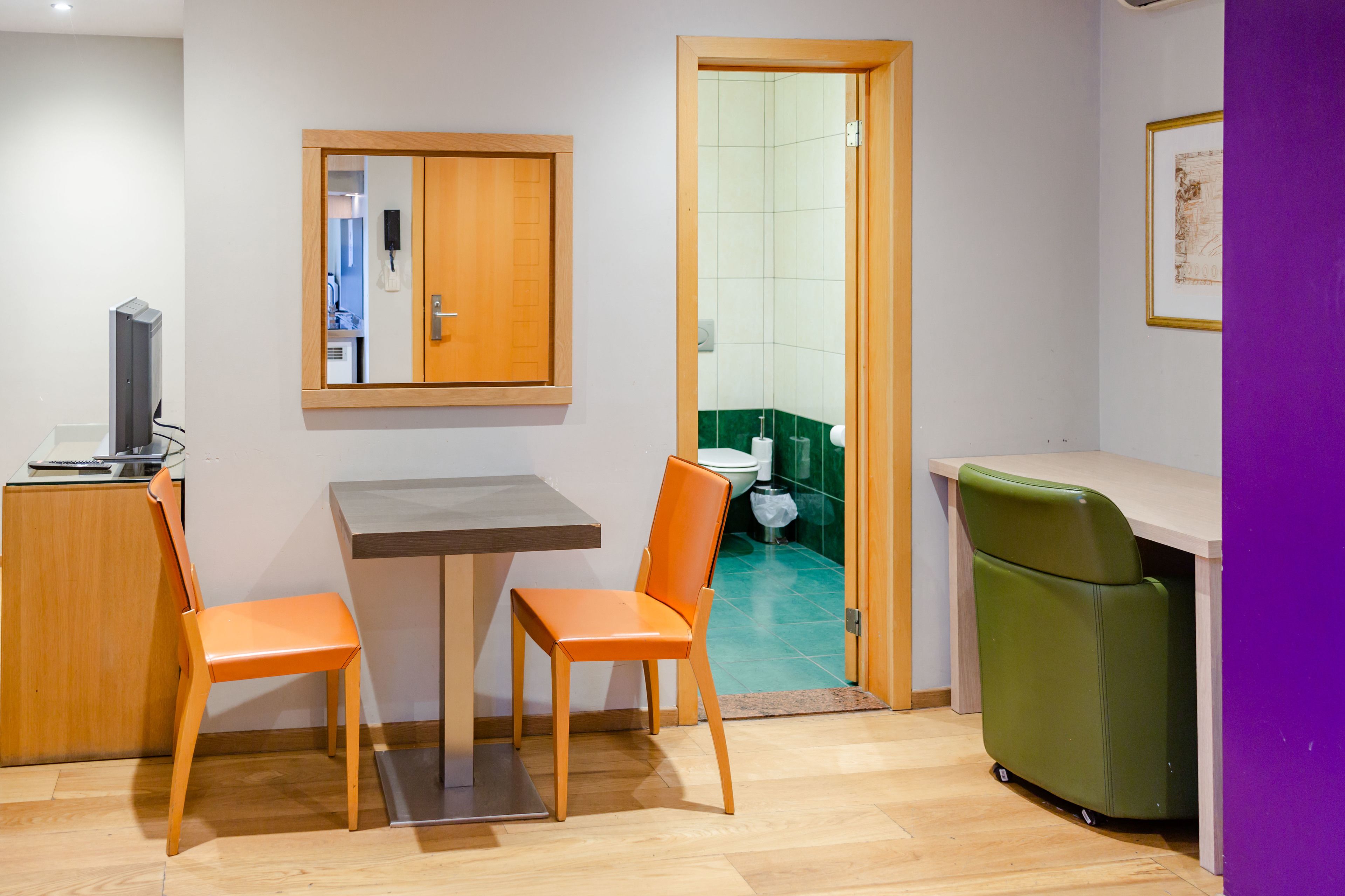 A small, modern room with a wooden table, two orange chairs, a green chair, a television, and a door leading to a bathroom.