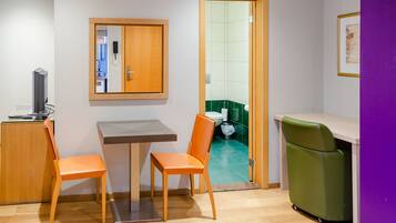 A small, modern room with a wooden table, two orange chairs, a green chair, a television, and a door leading to a bathroom.
