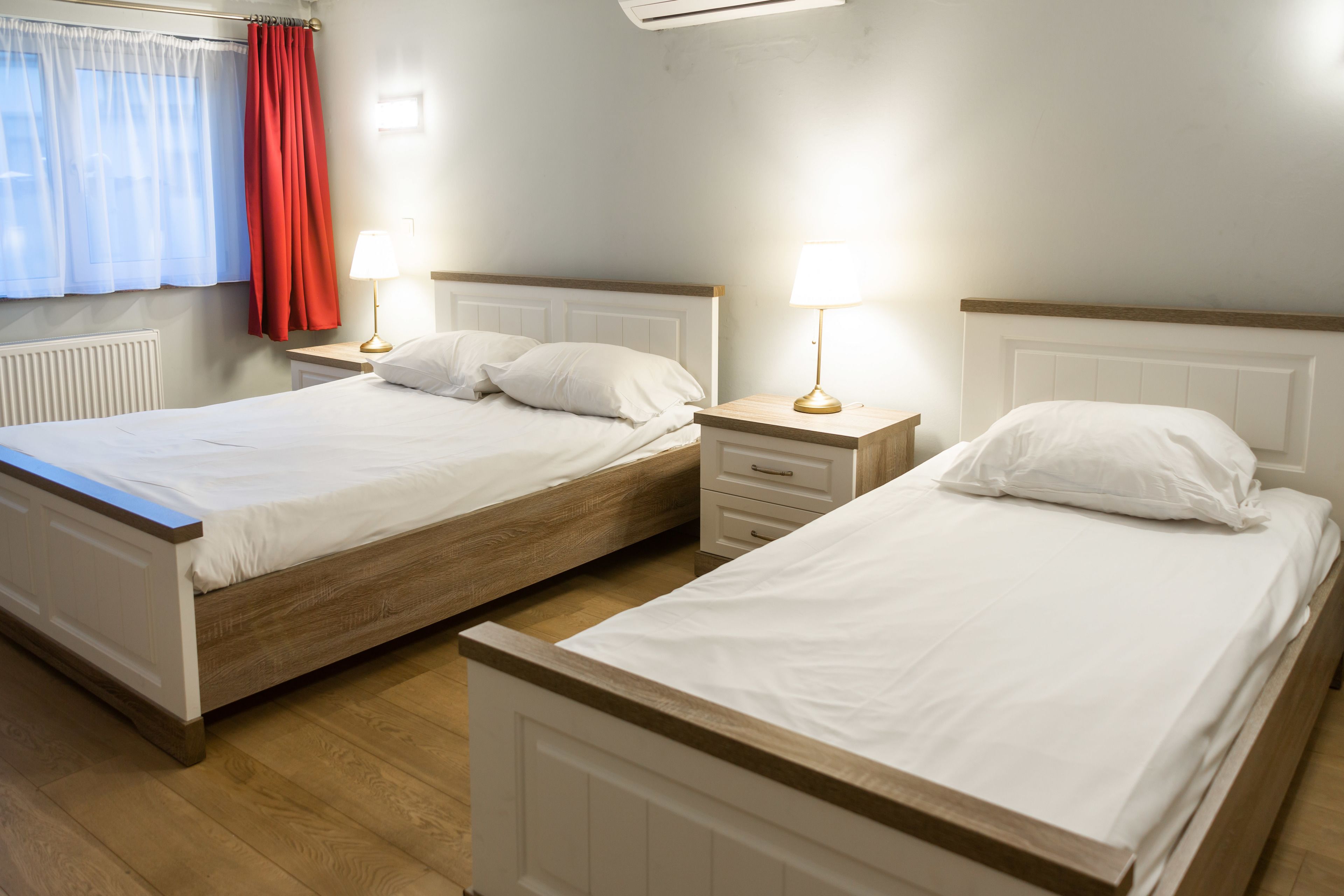 Et hotelværelse med to enkeltsenge, trægulv, en radiator og et vindue med gardiner.