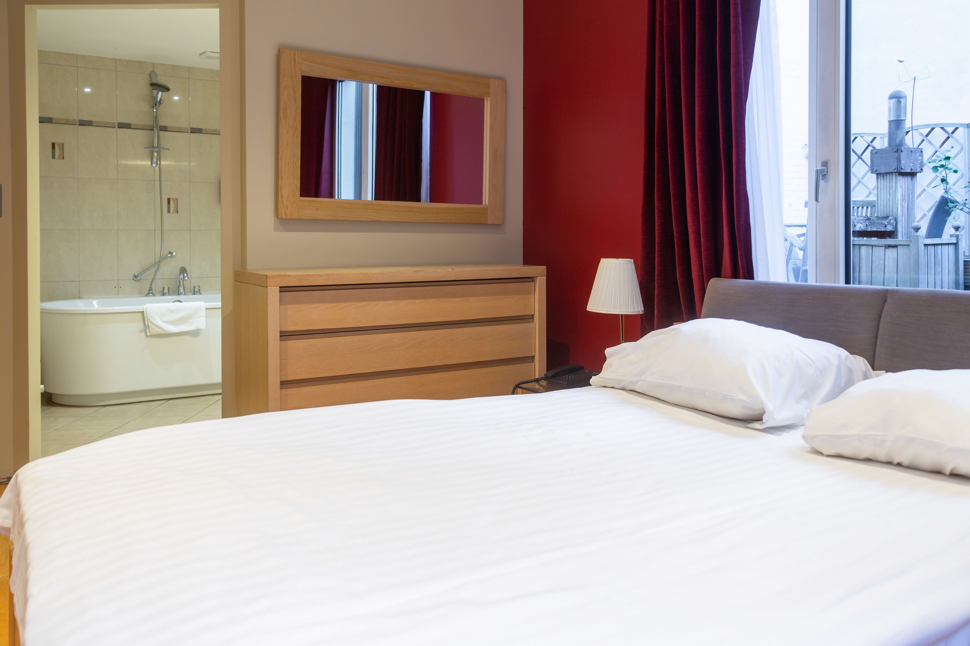 A hotel room with a bed, a wooden dresser, a mirror, a bathroom with a shower, and a view of a balcony.