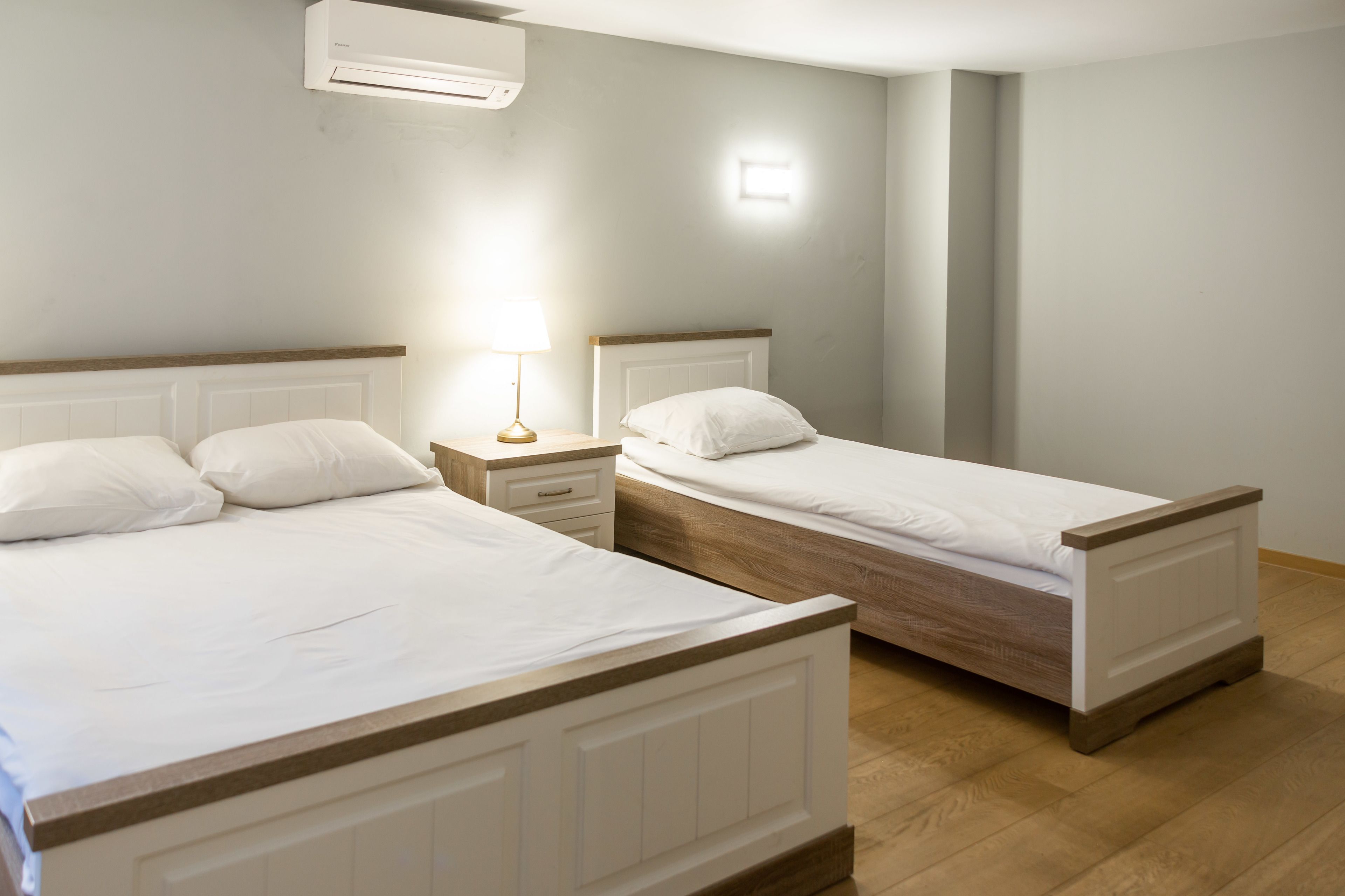 Two single beds with wooden frames and white bedding, a bedside table with a lamp, and a wall-mounted air conditioner.