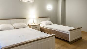 Two single beds with wooden frames and white bedding, a bedside table with a lamp, and a wall-mounted air conditioner.