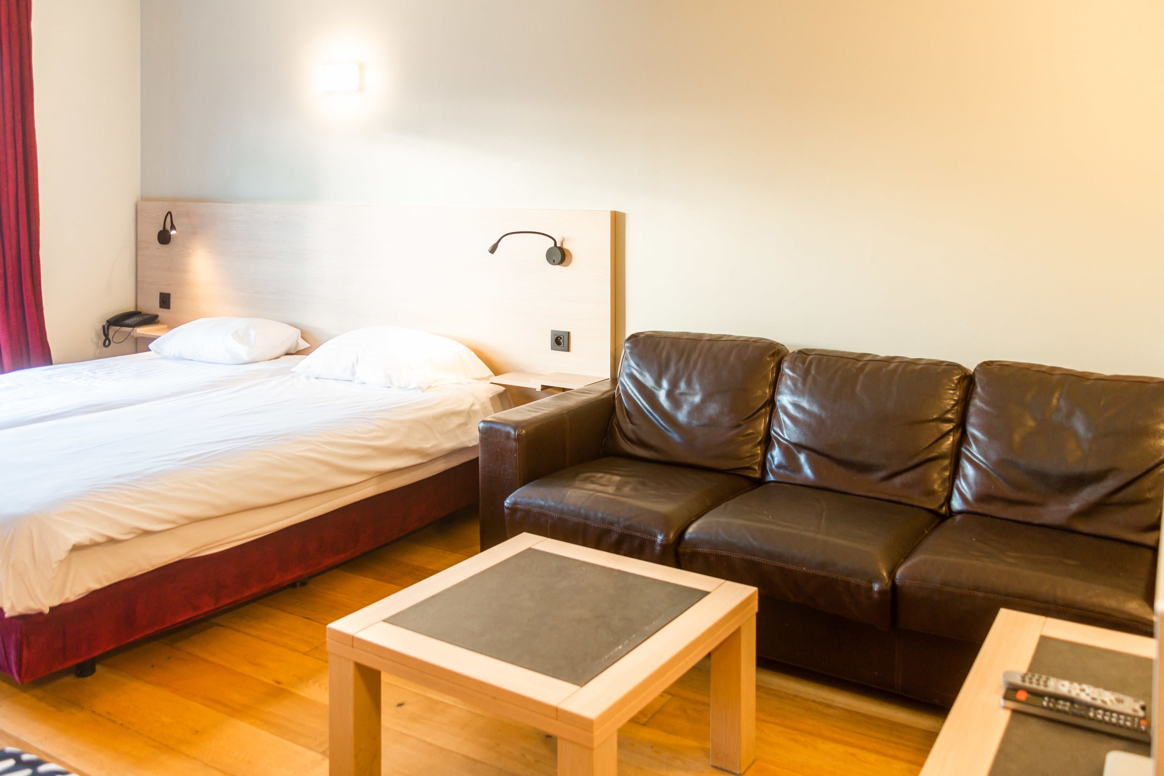 Quarto de hotel com cama, sofá de couro marrom, mesa de madeira pequena e luminária fixada na parede.
