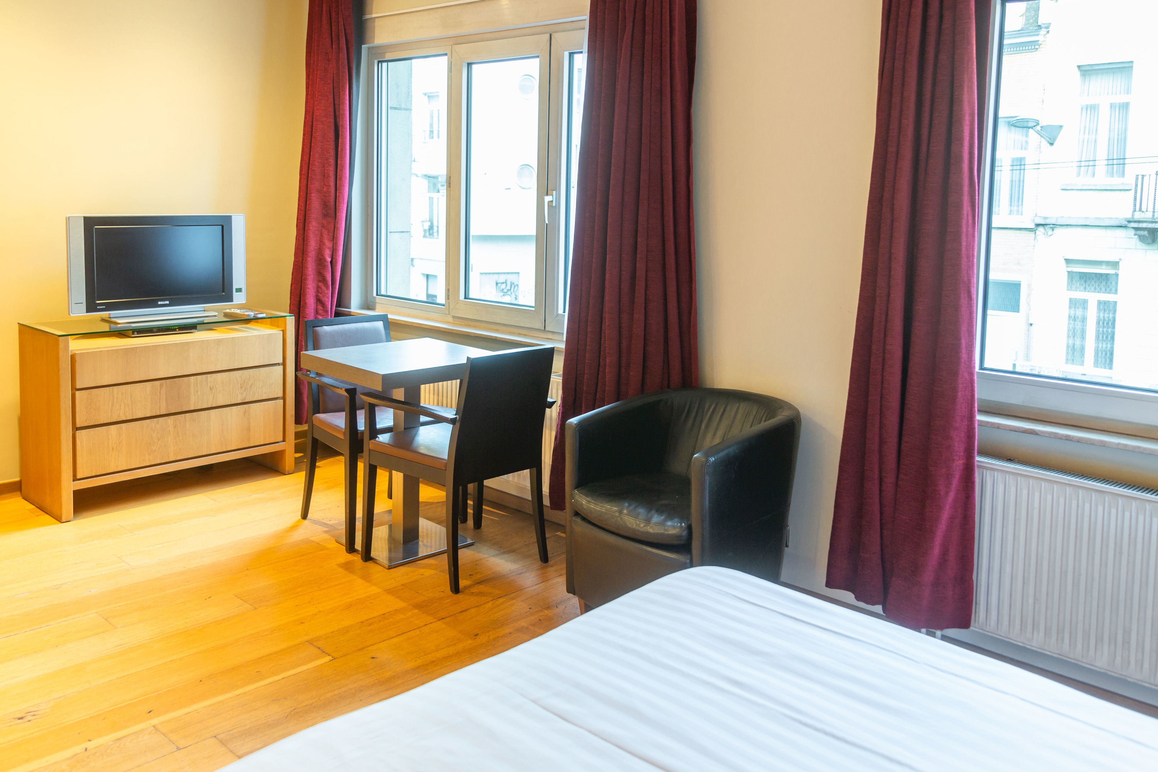 A hotel room with a bed, a desk with a chair, a television on a wooden cabinet, and a window with curtains.