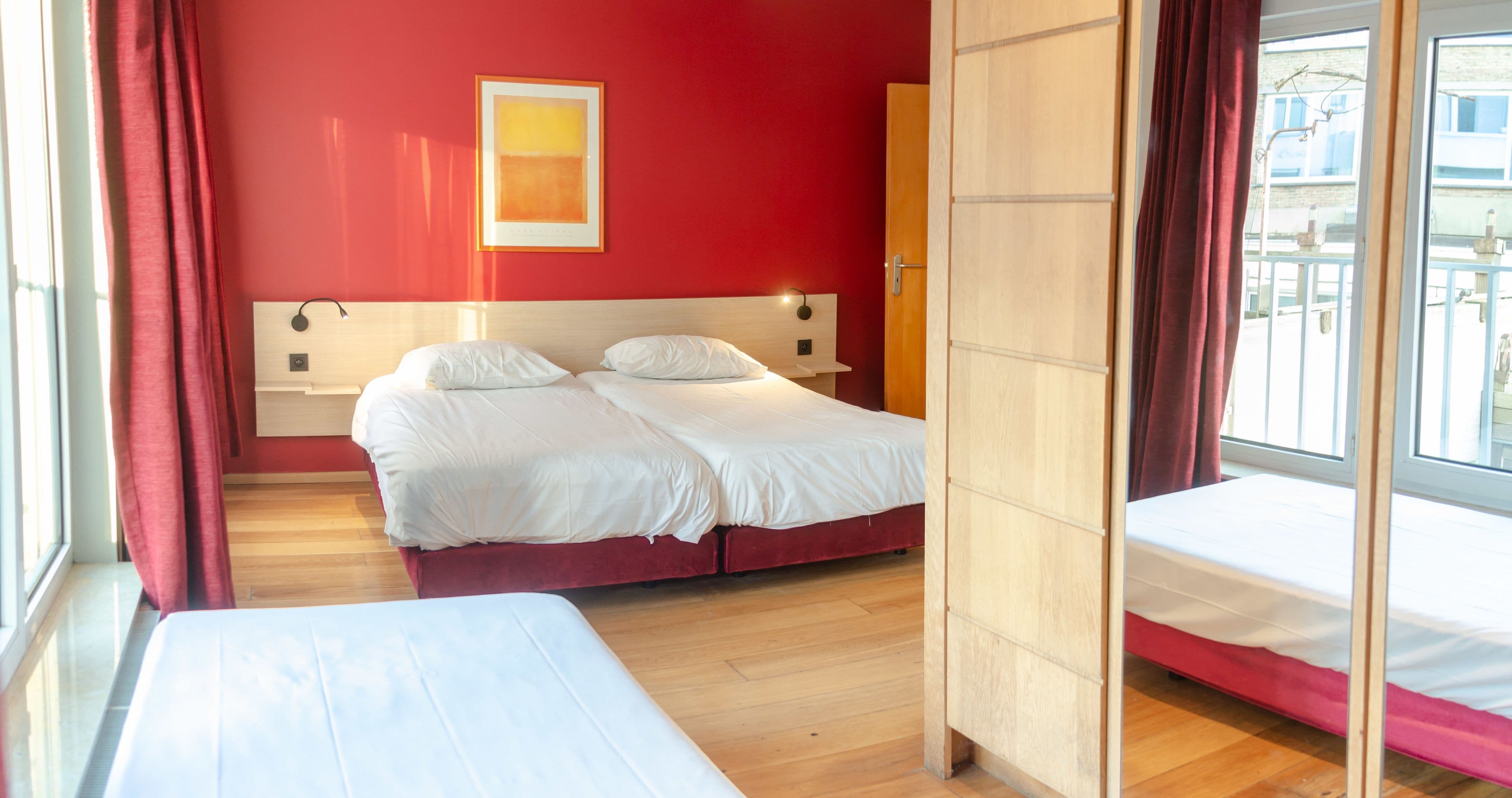 A hotel room with two beds, a red wall, and a window with curtains.