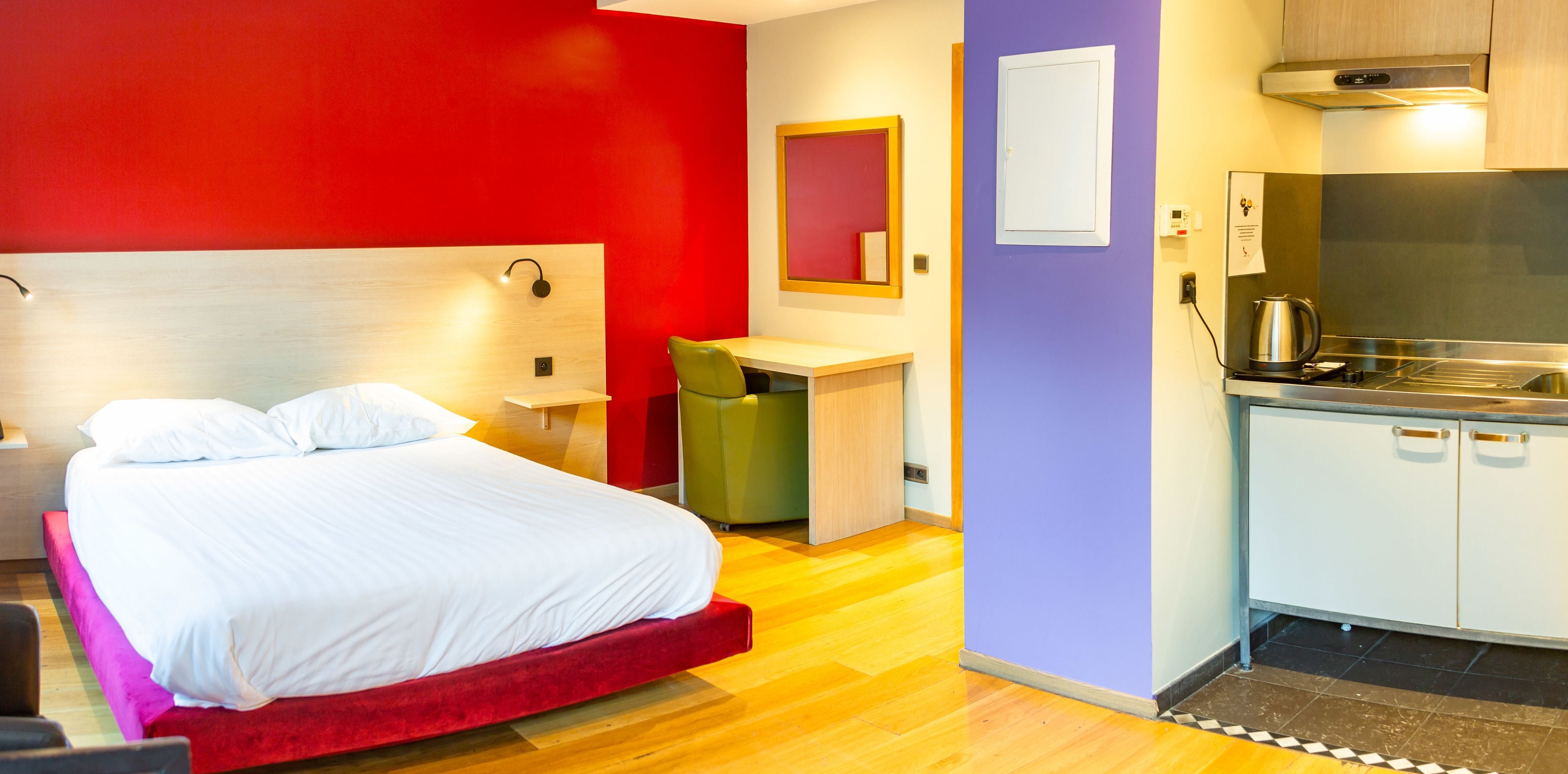 Una habitación de hotel con cama, escritorio, silla, kitchenette y espejo.