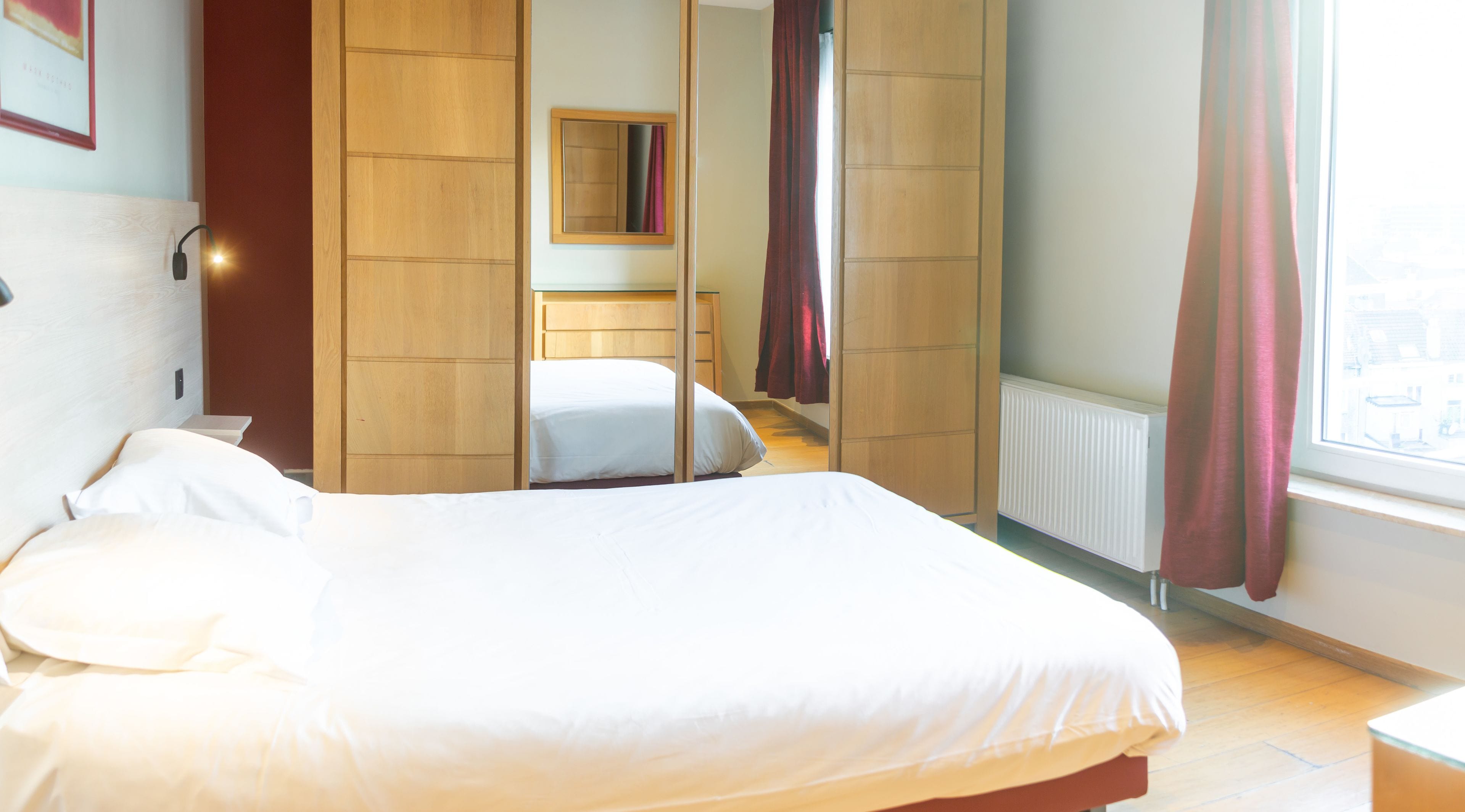 A hotel room with a bed, wooden wardrobe, a mirror, and a window with curtains.