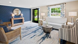 A modern living room with a sofa, two armchairs, a coffee table, and a side table. There is a large mirror on the wall, a window with curtains, and a door leading outside. - Love Farms Lodge (West Dennis)